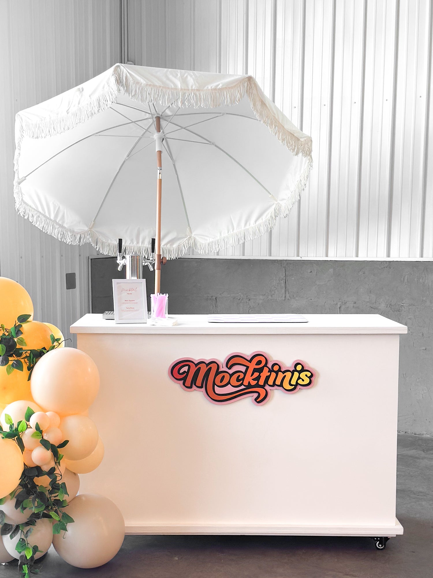 White mocktail tap bar with 'Mocktinis' branding under a white umbrella, surrounded by balloons and plants.