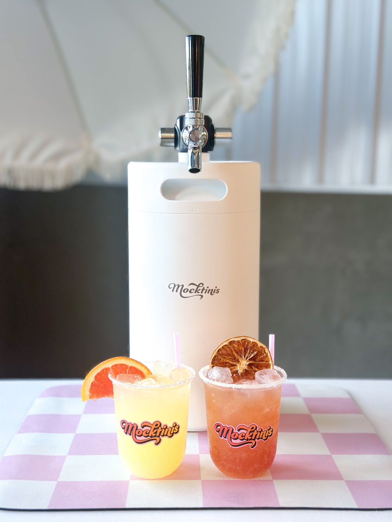 Two drinks with orange slices in front of a 'Tini Tap on a checkered serving mat.